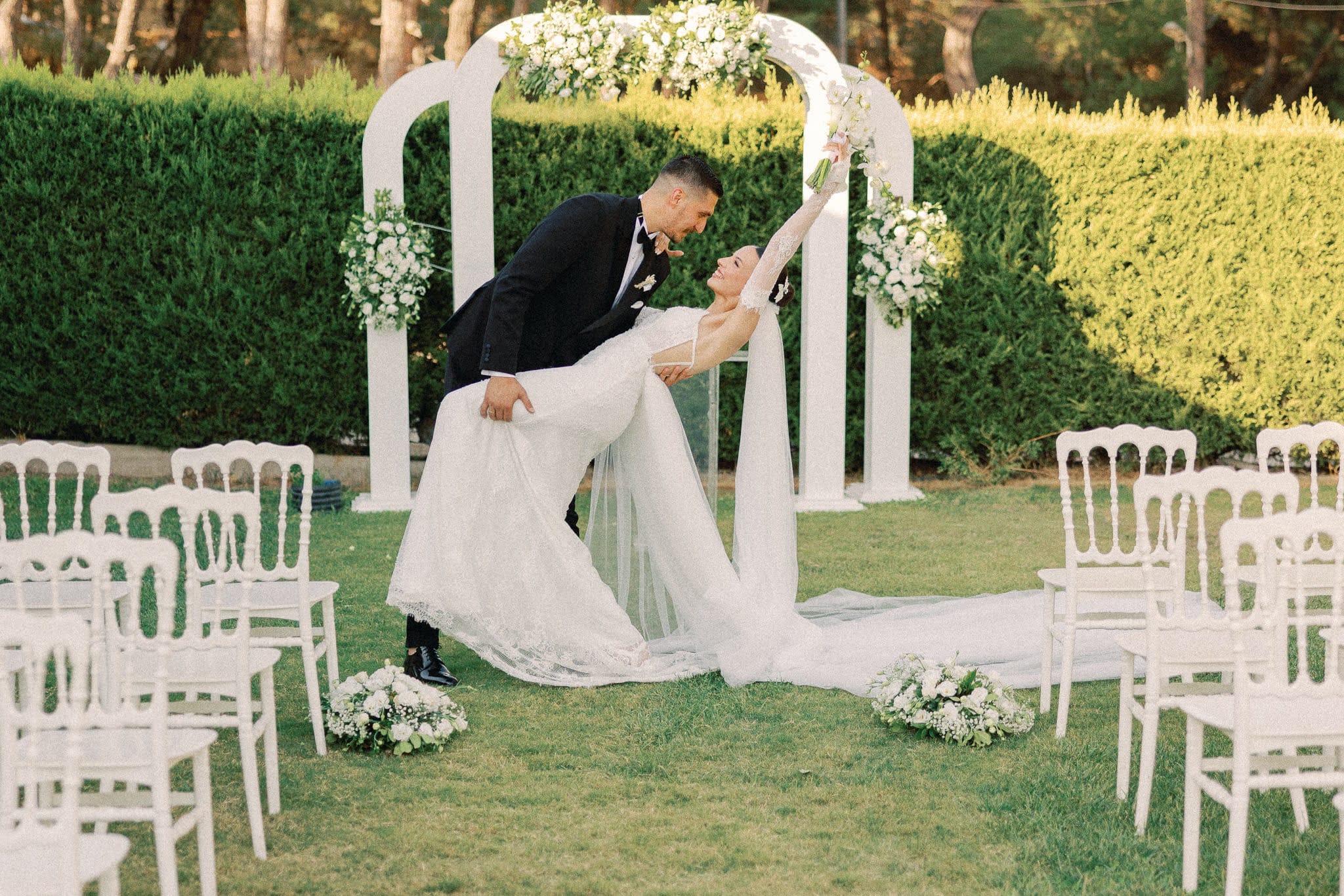 Beyaz sandalyeler arasındaki çiçekli tören kemerinin önünde dans pozu veren gelin ve damat